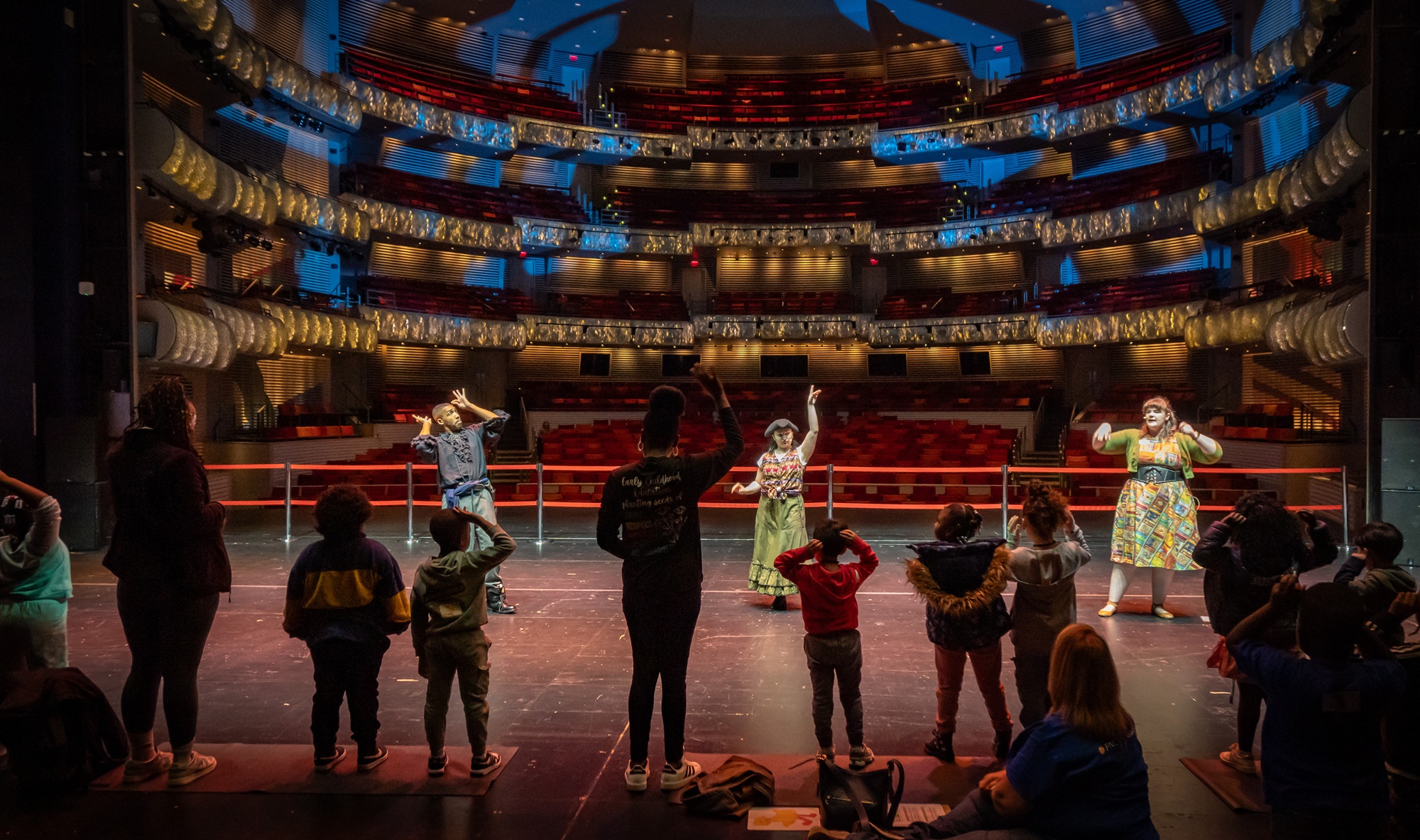 A group of children and adults stand on a theater stage, facing three performers demonstrating movements, with empty seats visible in the brightly lit auditorium.