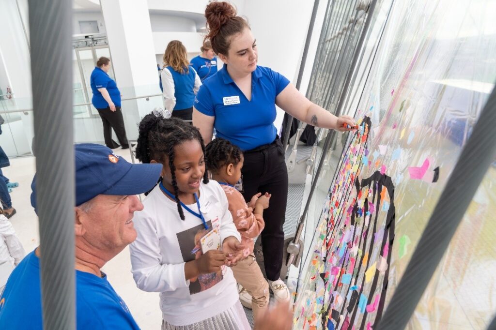 A group of people, including children and adults, place colorful stickers on a large transparent wall in a bright indoor setting.