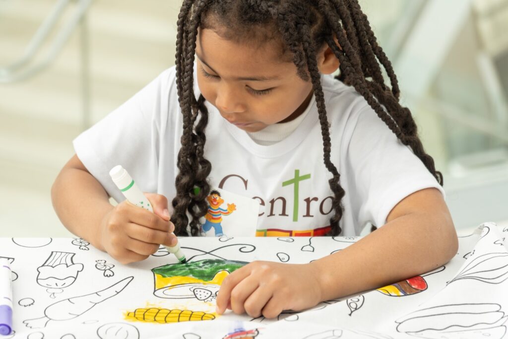 A child with braided hair colors a food-themed drawing on fabric using green and yellow markers.