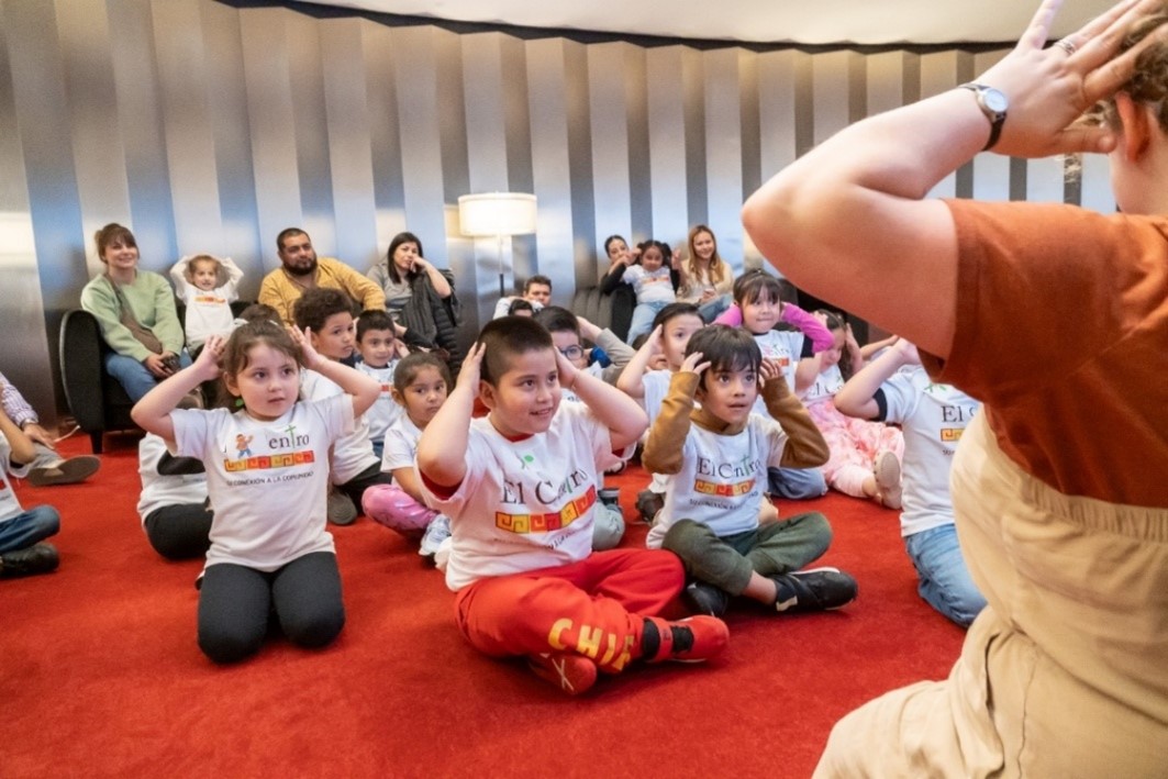 A group of children sit on a red carpet, following a seated adult’s instructions with their hands on their heads, while adults watch from chairs in the background.