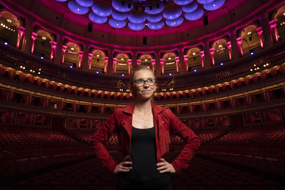 A woman in a red jacket stands confidently in the center of an ornate, empty theater with dramatic pink and purple lighting.