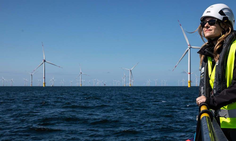 A woman in safety gear stands on a platform overlooking the ocean, with multiple offshore wind turbines visible in the background under a clear blue sky.