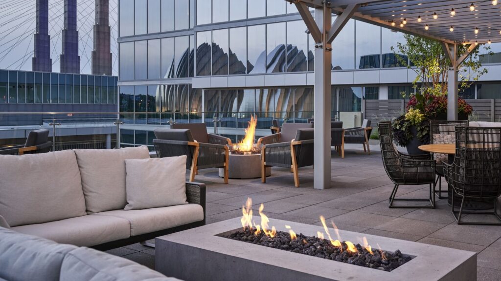 The modern rooftop terrace Of Loews Kansas City Hotel with cushioned seating, fire pits, and string lights; city buildings, including the Kauffman Center, are visible in the hotel's glass windows.