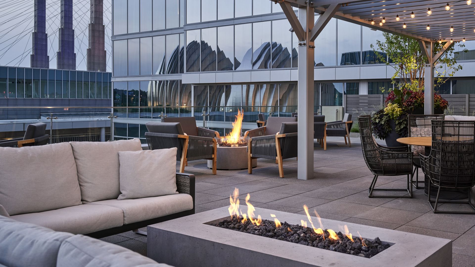 The modern rooftop terrace Of Loews Kansas City Hotel with cushioned seating, fire pits, and string lights; city buildings, including the Kauffman Center, are visible in the hotel's glass windows.
