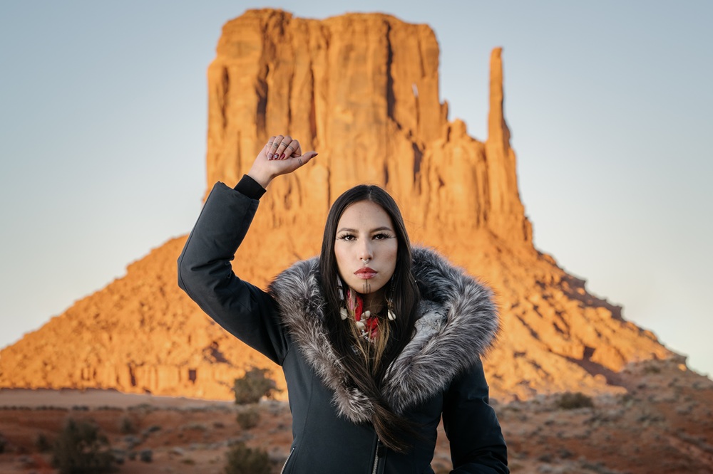 Portrait of Quannah Rose Chasinghorse-Potts, in front of sacred Indigenous land near her childhood home on the Navajo Nation. The landform is known as West Mitten, in Monument Valley, Utah. Set aside in 1958, it is a Navajo Nation tribal park, considered sacred by Diné and managed by the nation. Self-management of lands sacred to Indigenous peoples is one major aspect of the Native American landback movement.