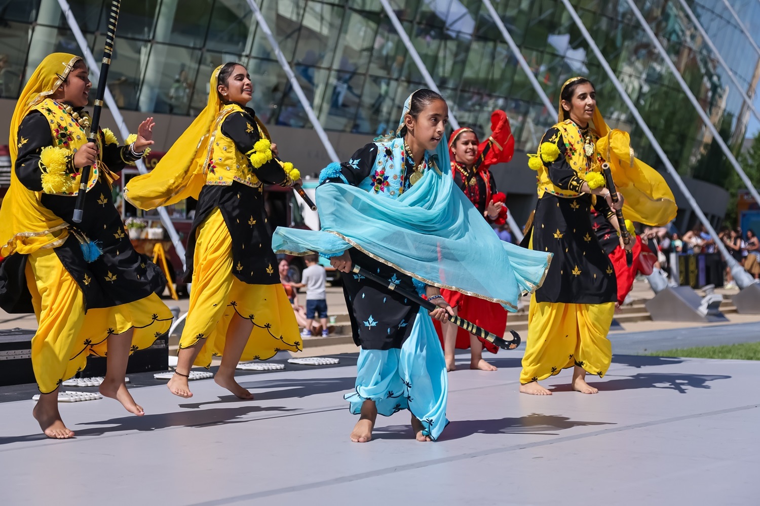 A group of girls in colorful attire perform a dance on an outdoor stage in front of the Kauffman Center, showcasing the vibrant spirit of the performing arts in Kansas City.