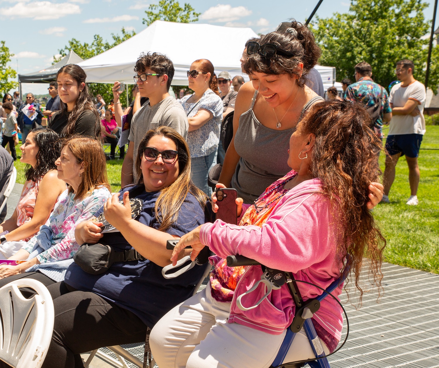 A group of people, including a woman in a mobility aid, gather outdoors at a sunny community event with tents and trees in the background.