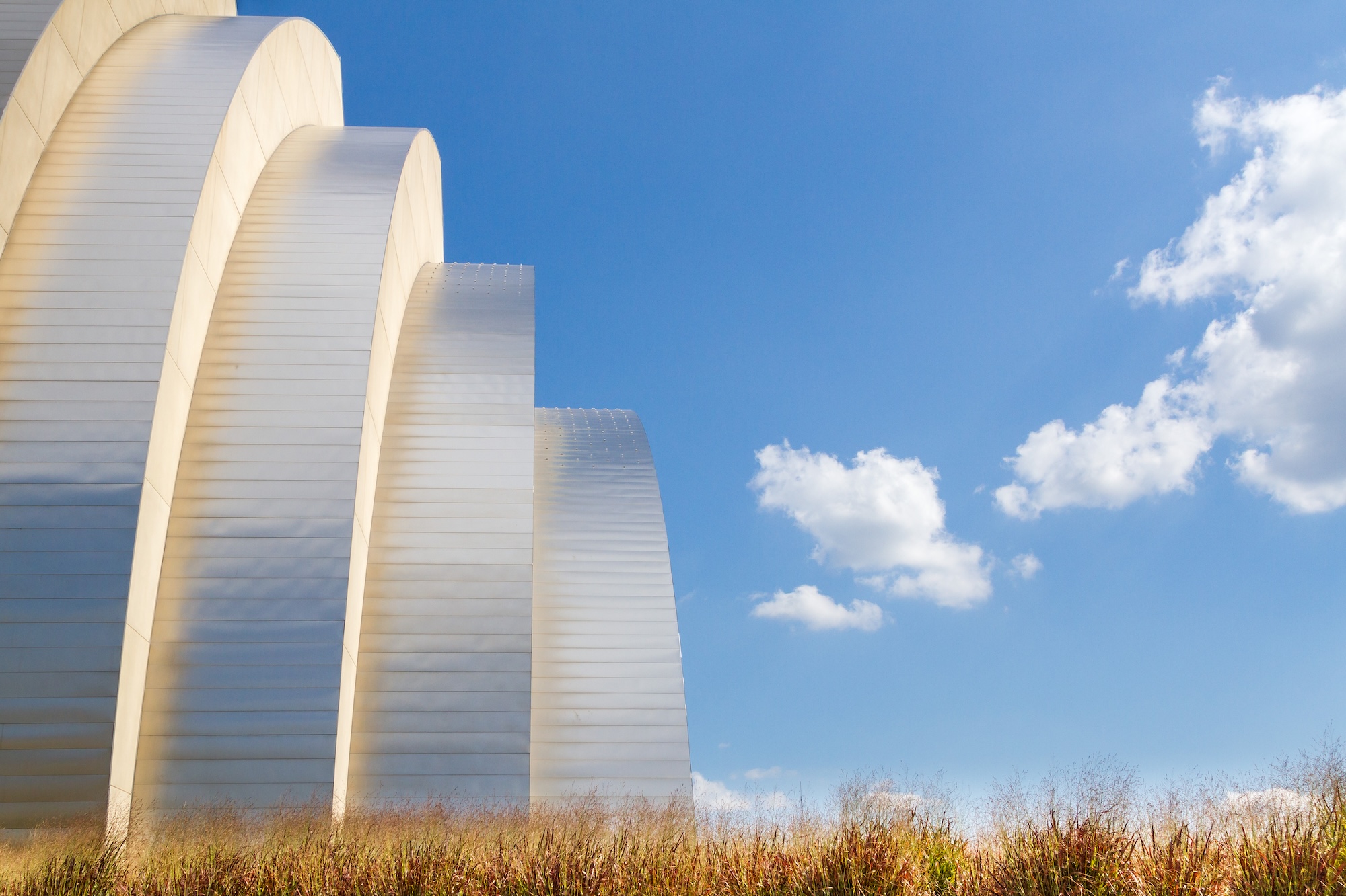 Curved, metallic building facade with layered arches stands against a blue sky with scattered clouds and dry grass in the foreground — the striking exterior of the Kauffman Center.